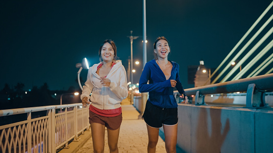 Multiracial team young sporty Asian women wear sports outfits running enjoy cardio through the city streets over bridge at night. Running club and exercise outdoor concept.