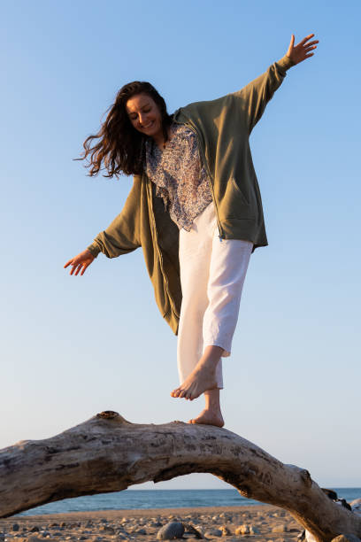 femme heureuse en équilibre sur un tronc d’arbre flotté à la plage au coucher du soleil dans l’heure dorée. jeune femme s’amusant à marcher sur un tronc d’arbre au bord de la mer. - driftwood wood water sunrise photos et images de collection