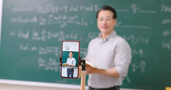 Asian senior male calculus professor is teaching with blackboard and having lecture online through smartphone at college classroom