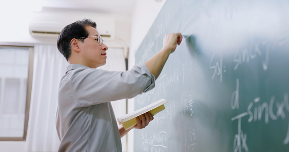 close up side view asian senior male calculus professor is writing in front of blackboard at college classroom