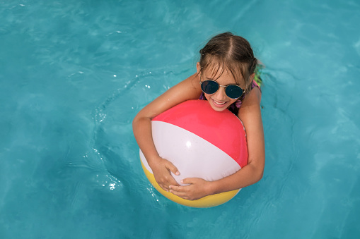 A cute happy laughing girl in blue sunglasses swims hugging an inflatable ball for a swimming pool.