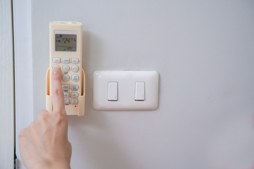 Index finger of woman hand touching at open button on air conditioner remote controller installing beside light switch at home with white wall copy space.