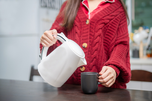 Crop Asian woman hand pouring hot water to black mug to make a cup of tea, person using energy by using boiling water pot with body of woman in red sweater background.
