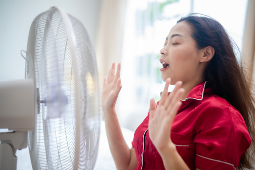 Asian beautiful woman in red and long hair standing in front of electrical fan, showing hands to touch the wind from fan with smile of happiness, freshness and relaxing emotion.