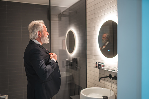 Mature businessman standing in front of a mirror in a bathroom dressing up. He is wearing a business suit.