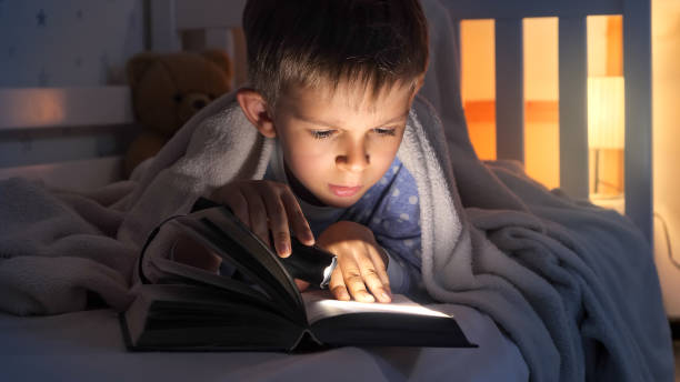 istock Cute boy in pajamas reading a book with flashlight in bed. Children education, development, secrecy, reading books. 1565519870