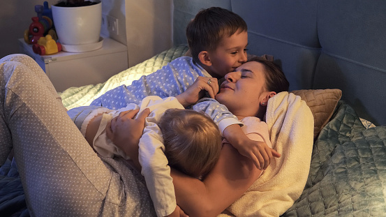 Two loving boys in pajamas hugging and kissing mother lying in bed at night. Family having time together, parenting, happy childhood and entertainment
