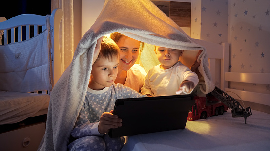 Happy family in bedroom at night watching cartoons on tablet computer. Family having time together, children with gadgets, parenting, happy childhood and entertainment