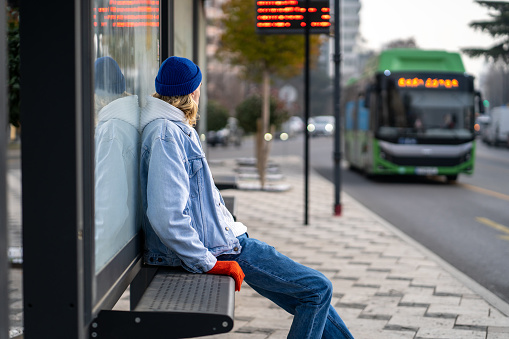 Millennial guy sitting on bench at bus stop looking at arriving bus, young man waiting at trolleybus station, people use public transportation to commute to work. Public transport timetable