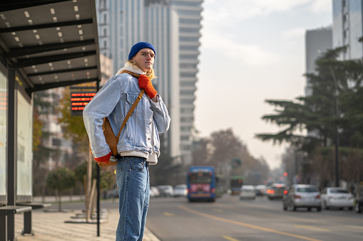 Caucasian guy student with backpack waiting for bus outdoors, young millennial man standing alone at public transport stop, living in big city. Transportation and new millennial generation, city life