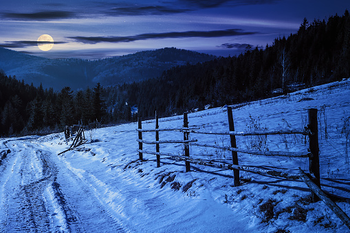 fence on the snow covered mountain slope near the forest in winter at night. countryside scenery in full moon light