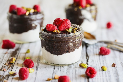 Dark chocolate chia pudding with raspberries and pistachios on top in a glass jar on a white wooden background. Served with coconut whipped cream. Healthy food. Copy space.