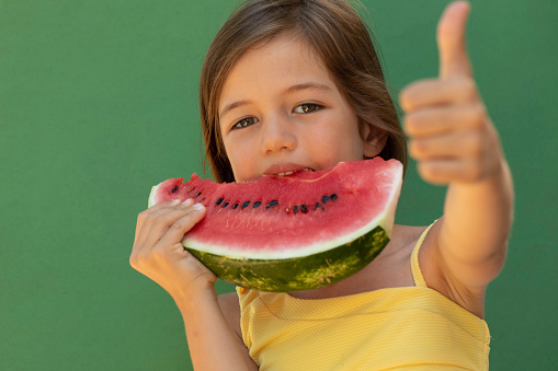 Girl eating a slice of watermelon in front of green background.