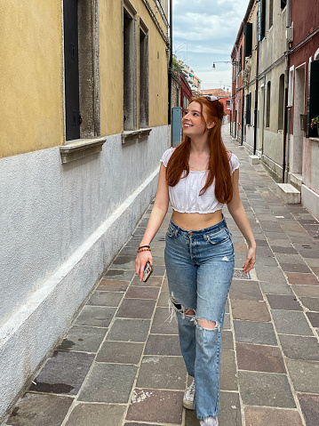 Stock photo showing close-up view of beautiful, red haired, female tourist wearing white crop top and ripped jeans whilst walking through Murano alleyways.
