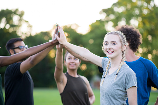 Smiling blonde young woman with her hair in braids and wearing athletic clothing is smiling at the camera while she is holding her hand up in a high five with her team in a group fitness class at a fitness club after they completed their exercises.