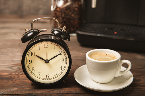 Alarm clock and a cup of morning coffee with a coffee machine on the background of the kitchen interior.A glass of coffee on the table with a clock in the morning. Good morning concept. WAKE UP!