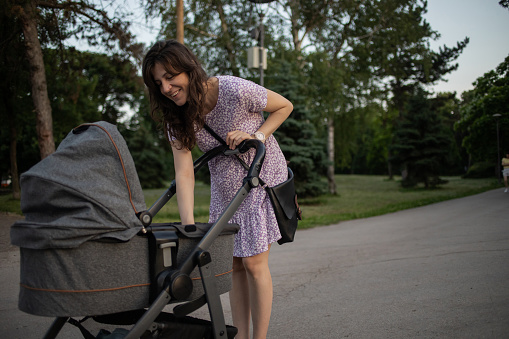 Pretty young woman walking through the public park with her baby son. He is in a baby carriage and this is their first walk.