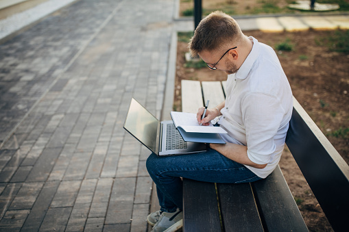 A young handsome businessman sitting on a bench outside and working on his laptop. He writes in his notebook.