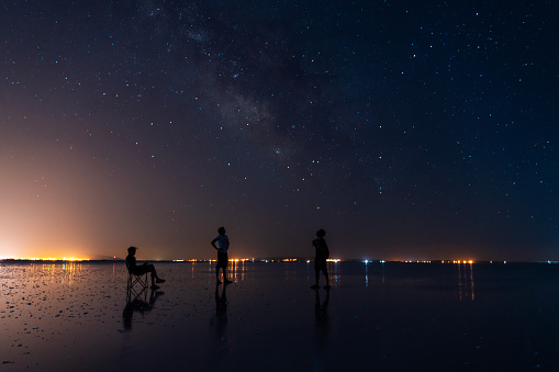 Young men looking at Milky Way view in Salt Lake. 
star reflection in still water. a starry dark night with no moonlight. The city lights are visible from far away. Long exposure with a full-frame camera at dusk.