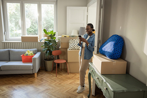Young woman moving in new home, unpacking boxes and enjoying in her new home. She is resting and using smart phone.