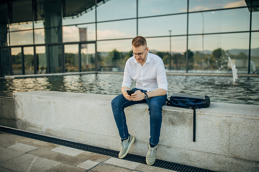 A young handsome man sitting outside near the fountain and using his mobile phone