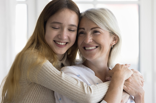 Cheerful teen granddaughter kid and happy grandmother enjoying warm family moment, meeting at home, hugging with toothy smiles and face touch, celebrating mothers day