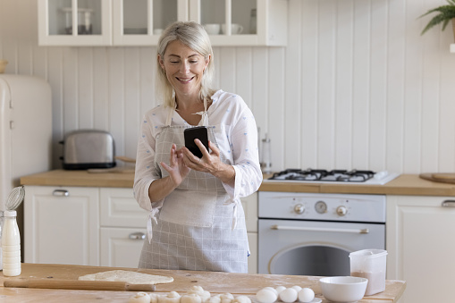 Cheerful baker blogger woman taking picture of baking process on smartphone for cooking blog, holding mobile phone over floury kitchen table with raw dough, roller, using Internet technology