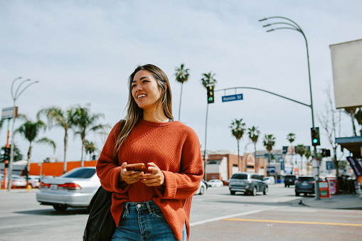 Stylish Asian - American woman walking on the streets of Los Angeles, California, in Van Nuys area of the Valley. She is using her smartphone, texting messages, checking apps while commuting to work.