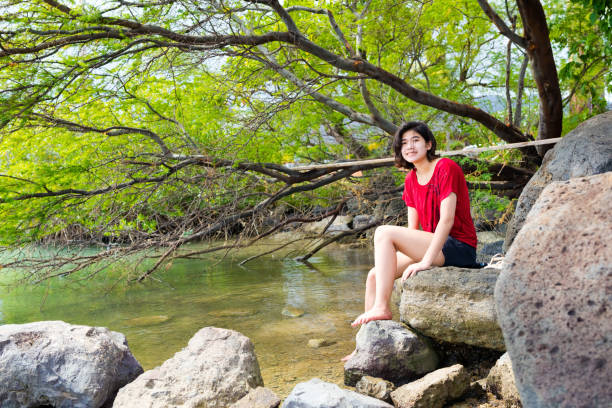 Young woman sitting and relaxing on large rocks in quiet bay off hawaiian ocean under shade trees