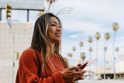 Stylish Asian - American woman walking on the streets of Los Angeles, California, in Van Nuys area of the Valley. She is using her smartphone, texting messages, checking apps while commuting to work.