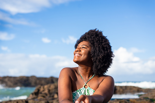 Portrait of a smiling woman wearing blue sitting on the rock of a beach looking to the side. Blue sky in the background. Rio Vermelho Beach, Salvador, Brazil.