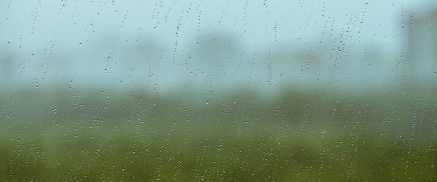 istock Atmospheric minimal backdrop with rain droplets on glass. Wet window with rainy drops and dirt spots closeup. Blurry buildings and green trees under blue sky against dirty window glass with raindrops. 1566097773