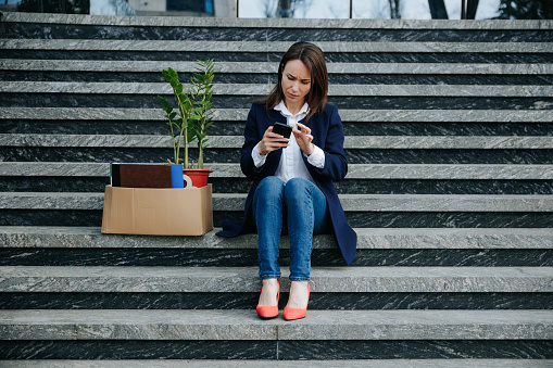 An Unhappy Woman, Recently Facing Dismissal, Sits on Staircase Searching for Job Opportunities on her Smartphone.