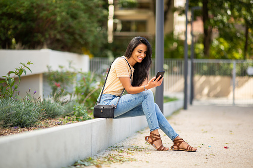 Full body portrait of young woman sitting outside looking at her mobile phone and smiling