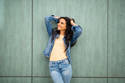 Portrait of fashionable young hispanic woman posing against green wall looking away with hands in hair