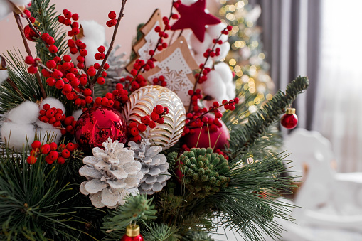 A bouquet of fir trees with red berries, cotton, toys on the table against the background of Christmas tree garlands. Winter decorations in the interior. The concept of preparation for the New Year holidays 2024.