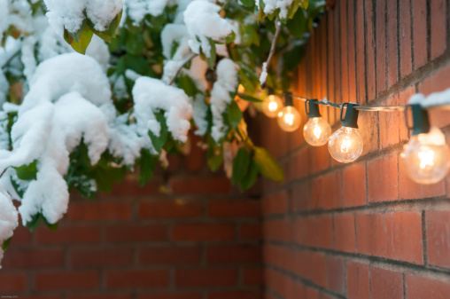 String of outdoor lights with a snow covered tree in background and brick wall bordering the image from a courtyard.