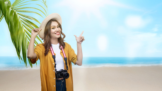 Asian woman with a hat and camera pointing at something while traveling on the beach with blue sky background