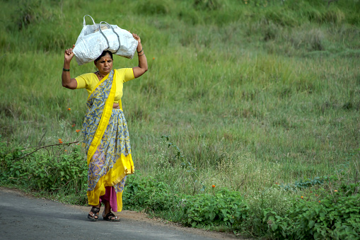 Pune, India - July 23 2023: Candid shot of an Indian lady walking while carrying a sack on her head at Uruli near Pune India.