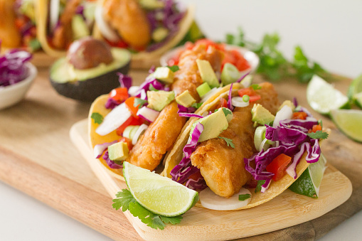 Beer Batter Fried Cod Fish Tacos, served with fresh shredded red cabbage, radishes, tomatoes, scallions, avocado, cilantro, and lime on a corn tortilla
