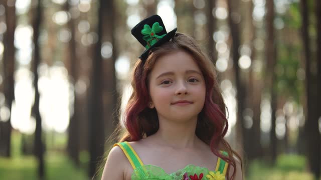 Cute leprechaun girl posing in the sunny forest. Beautiful little girl wearing green fairy dress and cylinder hat for a St Patricks Day celebration