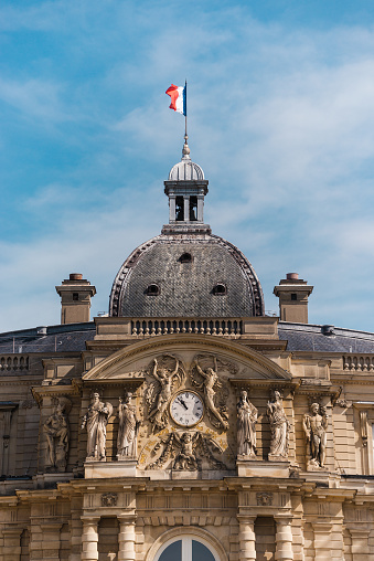 Paris, France - May 21, 2023: Clock tower detail of the Luxembourg Palace. The Jardin du Luxembourg (known in English as the Luxembourg Garden, or Senate Garden) is located in the 6th arrondissement of Paris, France. Creation of the garden began in 1612 when Marie de' Medici, the widow of King Henry IV, constructed the Luxembourg Palace as her new residence. The garden today is owned by the French Senate, which meets in the Palace. It covers 23 hectares and is known for its lawns, tree-lined promenades, tennis courts, flowerbeds, as well as picturesque Medici Fountain, built in 1620.
