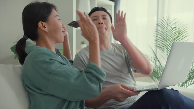Happy Asian couple having fun using laptop devices shopping use credit card online payment together sit on sofa in living room at home. Lifestyle spend time in house.