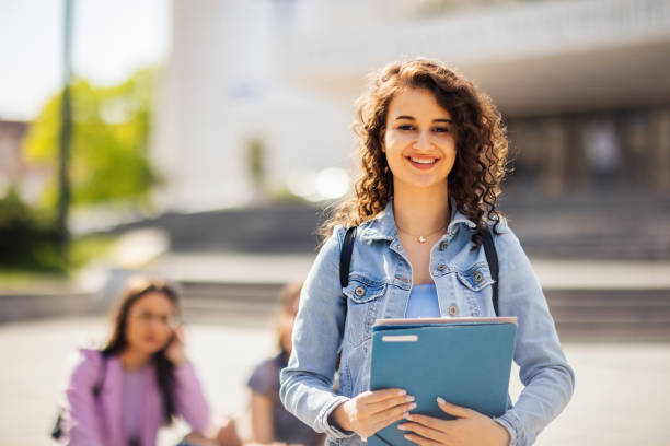 istock Student standing with her note-book while her friends are studying behind her 1566610109