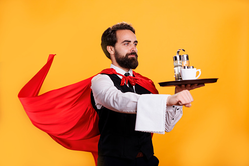 Superhuman butler with cape and platter, serving food at five star restaurant on camera. Young adult carrying tray with cutlery, posing as a fantasy superhero with red mantle in studio.