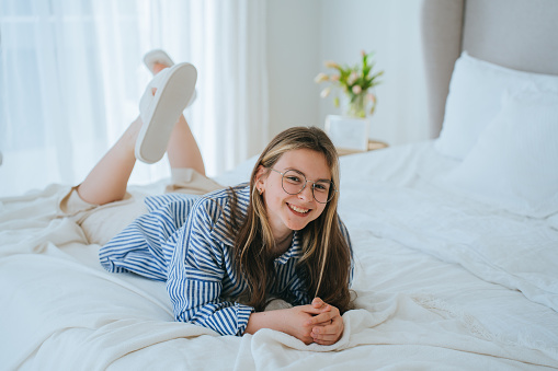 Young girl in casual laying on bed looks at camera smiling broadly enjoying vacations. Cheerful American schoolgirl relaxing at home. Domestic leisure. Teenagers on holidays.