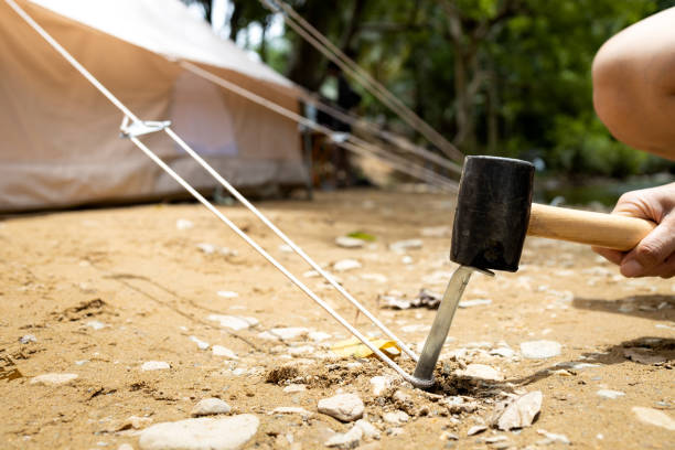 mão do macho segurando um martelo de borracha, martelando o aço do aço estaca estacas prego, tenda de fixação para acampar, bater ou bater âncora da barraca no chão, martelo com cabeça de borracha, punho de madeira - rubber mallet - fotografias e filmes do acervo