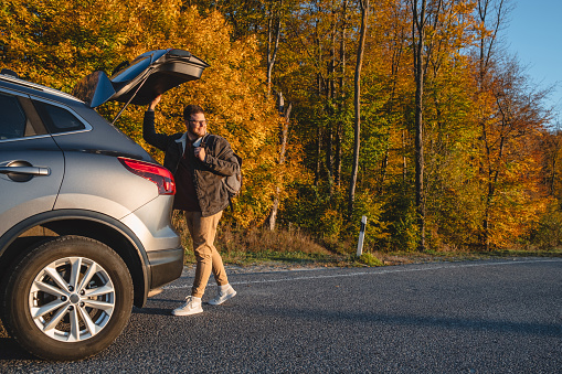 smiling man with backpack on his back closes trunk of car in autumn season. copy space