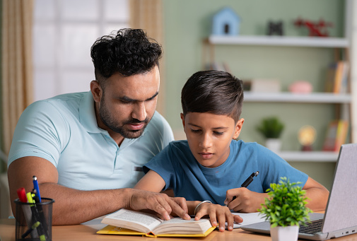 Indian father teaching or helping the son for reading from book at home - concept of Parental Guidance, home education and Tutoring Sessions