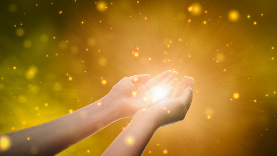 Conceptual Visualization Of Male Or Female Hands Reaching Out In Prayer On Magical Dark Yellow Background. Person Connecting With Higher Spiritual Energy Through Bright Light In Their Palms.